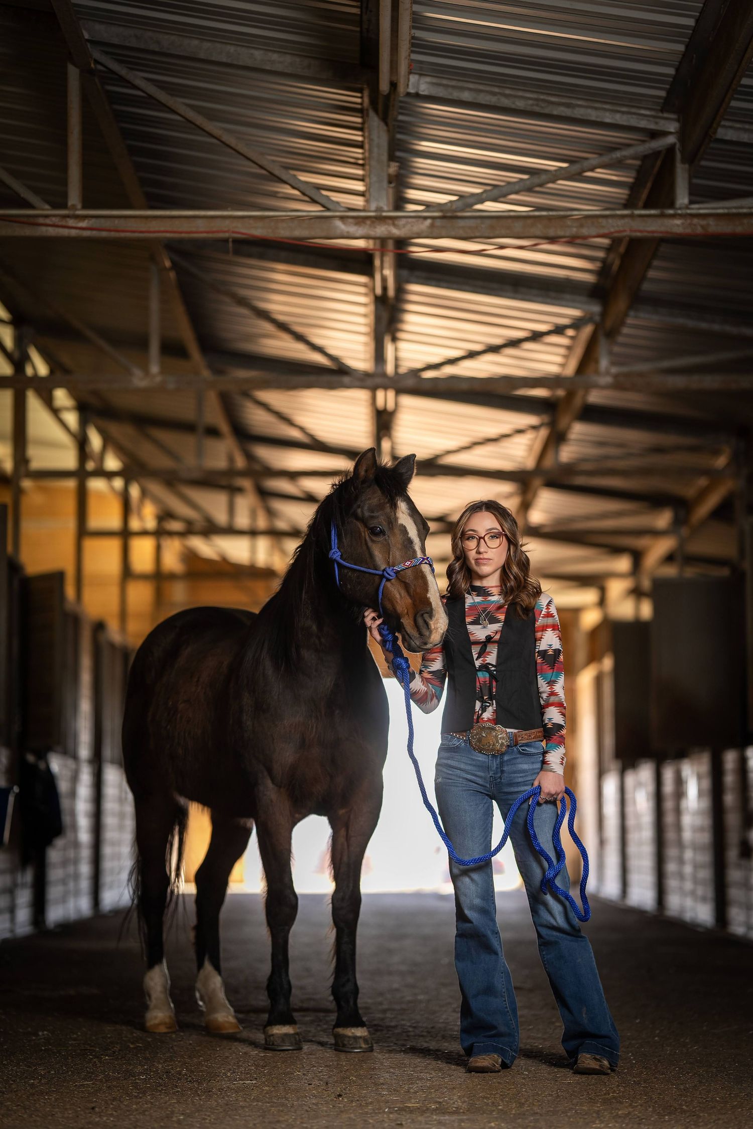 A dark horse and person stand together in a covered stable walkway with dramatic lighting and shadows.