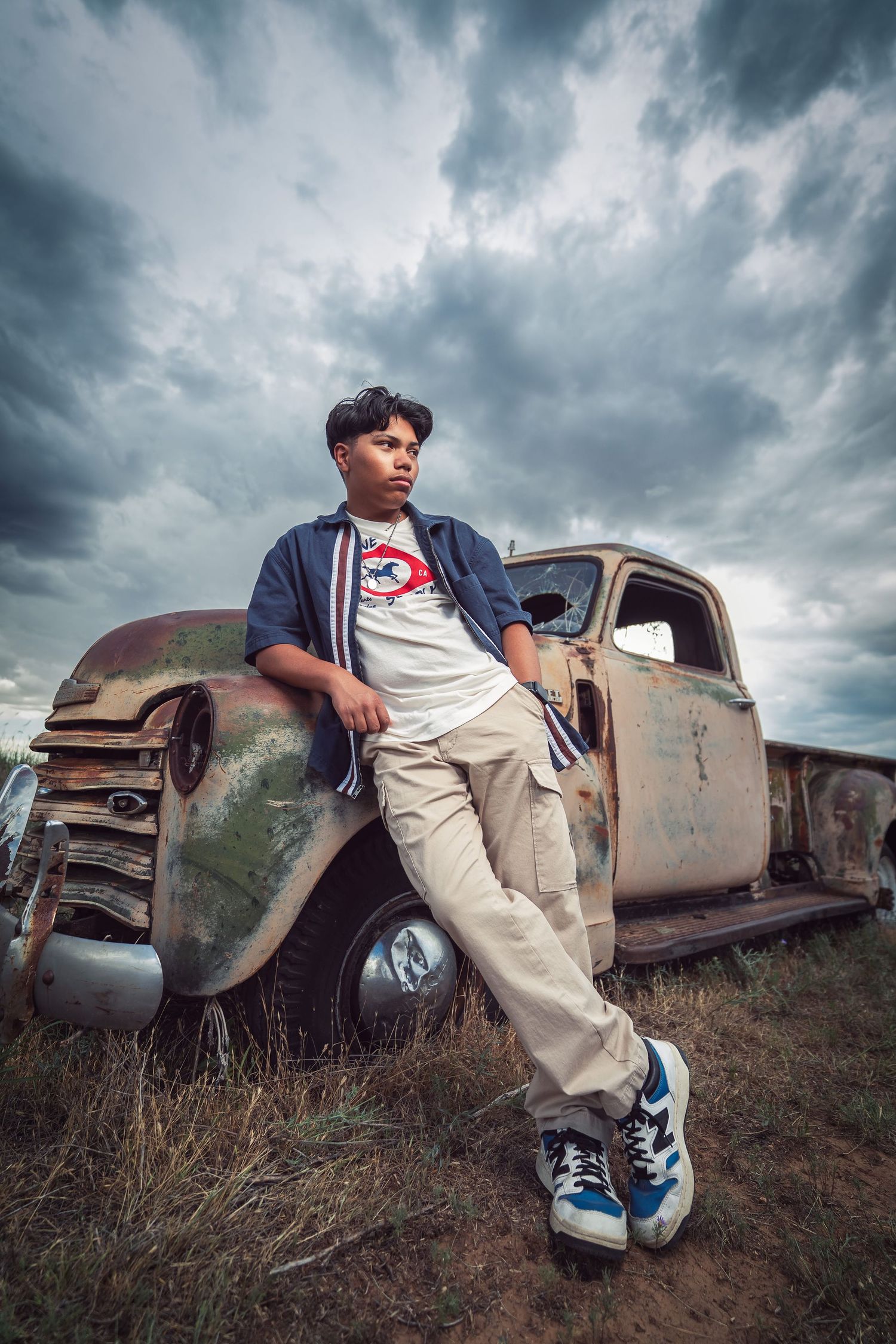 A person sits on a rusty vintage car in an overgrown field under dramatic stormy clouds.