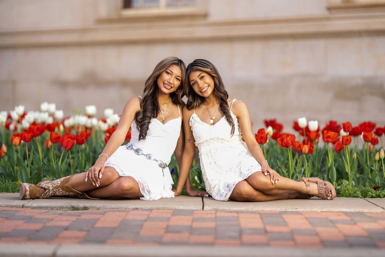 Two people in white dresses lay together on grass near red tulips and brick pavers in a garden setting.