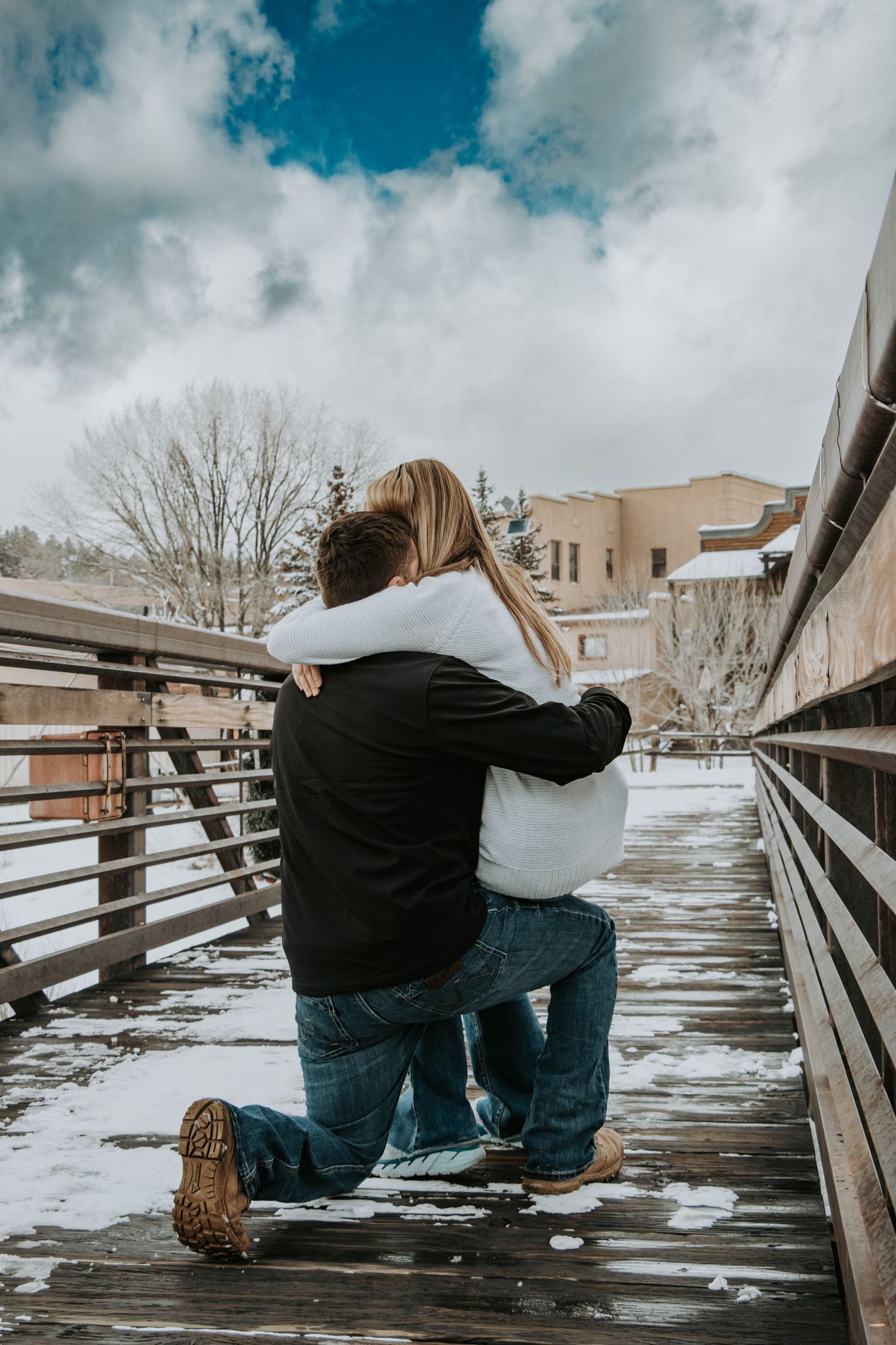 A romantic proposal moment on a snow-covered wooden bridge under cloudy skies.