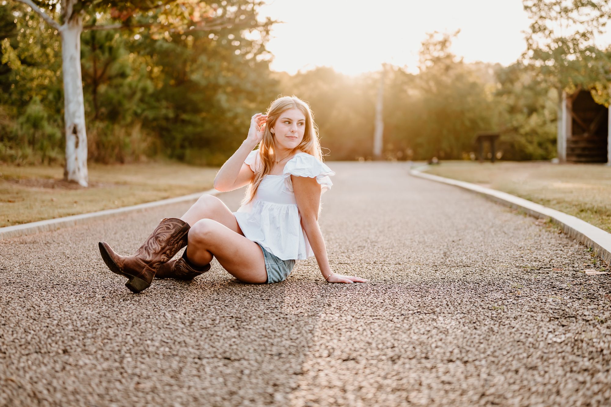 Young woman in white top, denim shorts, and cowboy boots sitting on a sunlit road surrounded by trees.