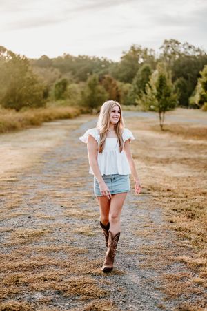 Young woman in white top, denim shorts, and cowboy boots walking on a gravel path at golden hour.