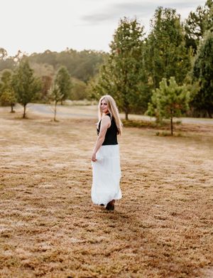 Young woman in black top and white maxi skirt walking through a dry grassy field with green trees behind her.