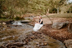 Woman in white skirt sits on rocks beside a peaceful woodland creek surrounded by autumn trees.