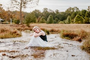 A woman in a white dress sits gracefully in a shallow muddy puddle in a rustic field with autumn trees.