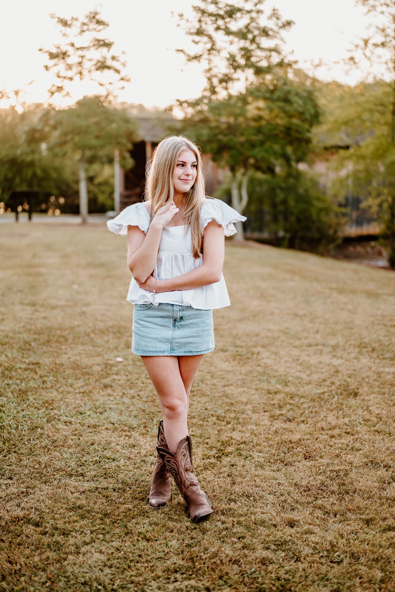 Young blonde woman in white ruffled top, denim mini skirt, and cowboy boots standing on green lawn at golden hour.