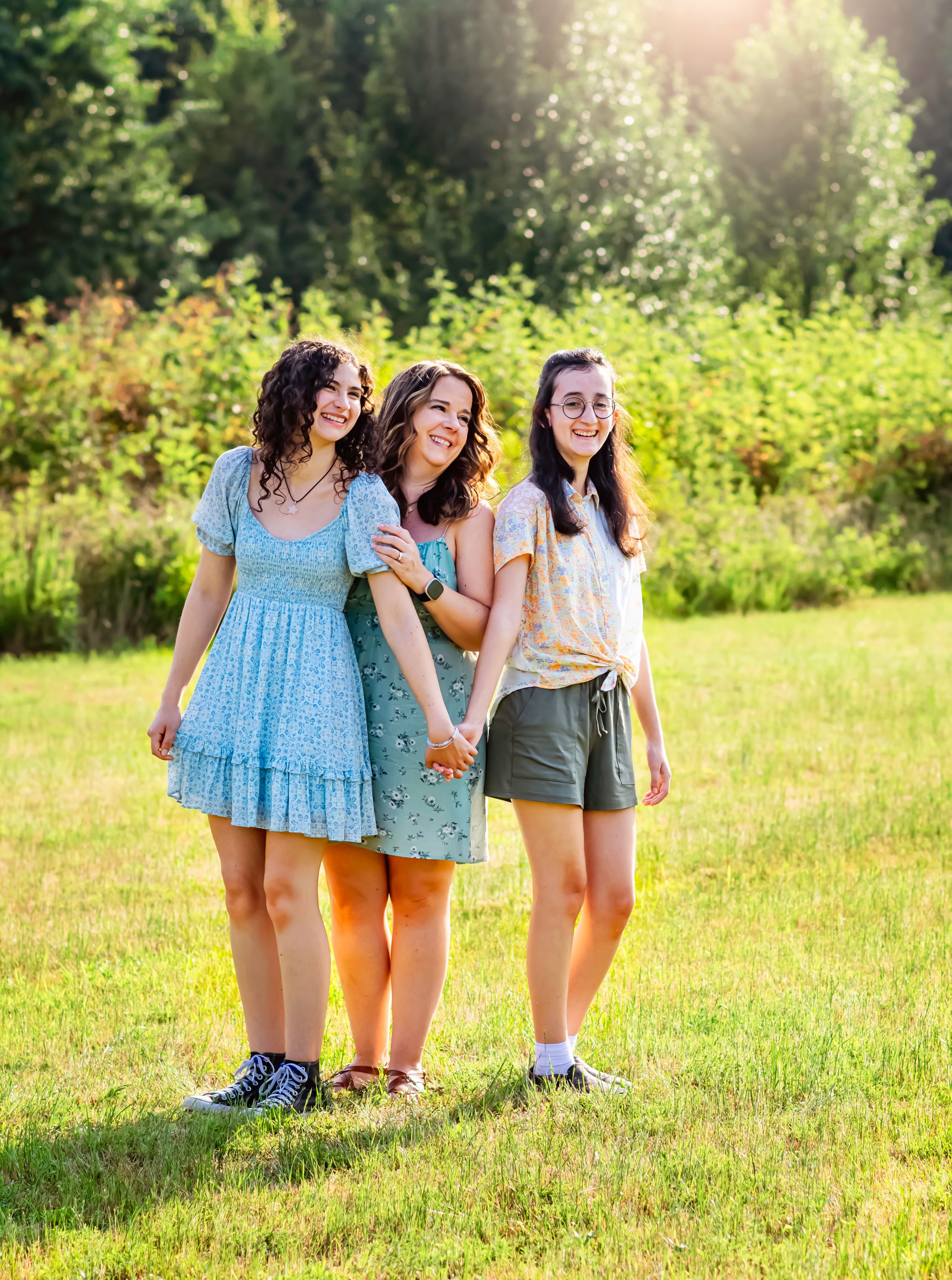Three friends in summer dresses laugh together in a sunlit meadow.