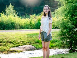A person in casual attire stands near a stream in a lush summer setting.