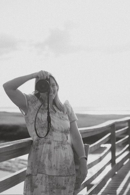 Woman in floral dress holding camera up to face on wooden boardwalk in black and white photo.