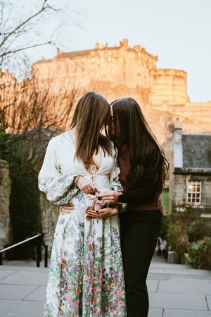 A couple shares an intimate embrace outdoors at sunset with stone buildings and trees in the background.