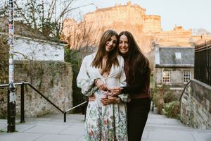 Two people share a cheerful moment together on stone steps with Edinburgh Castle visible in the warm sunset backdrop.