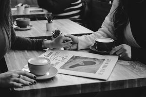 Black and white photo shows hands writing next to coffee cups and papers on a table in natural light.