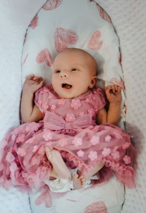 Happy baby wearing a pink polka dot dress lying on white fabric with heart patterns.