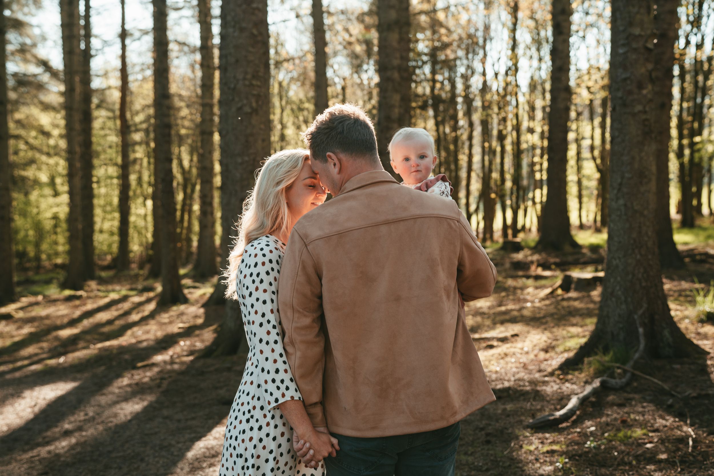 Couple walks hand in hand through a sunlit forest with golden rays streaming through tall pine trees.