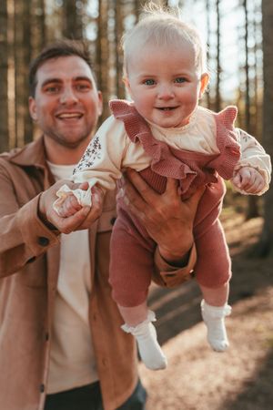 Adorable toddler being held up in playful poses outside in autumn forest with warm brown tones.