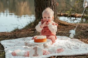 A joyful baby wearing a red dress plays in white cake frosting and smears it during their first birthday photoshoot.