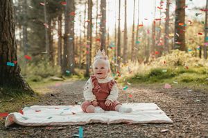 Small child in red overalls sits on blanket in forest path while autumn leaves fall around them.