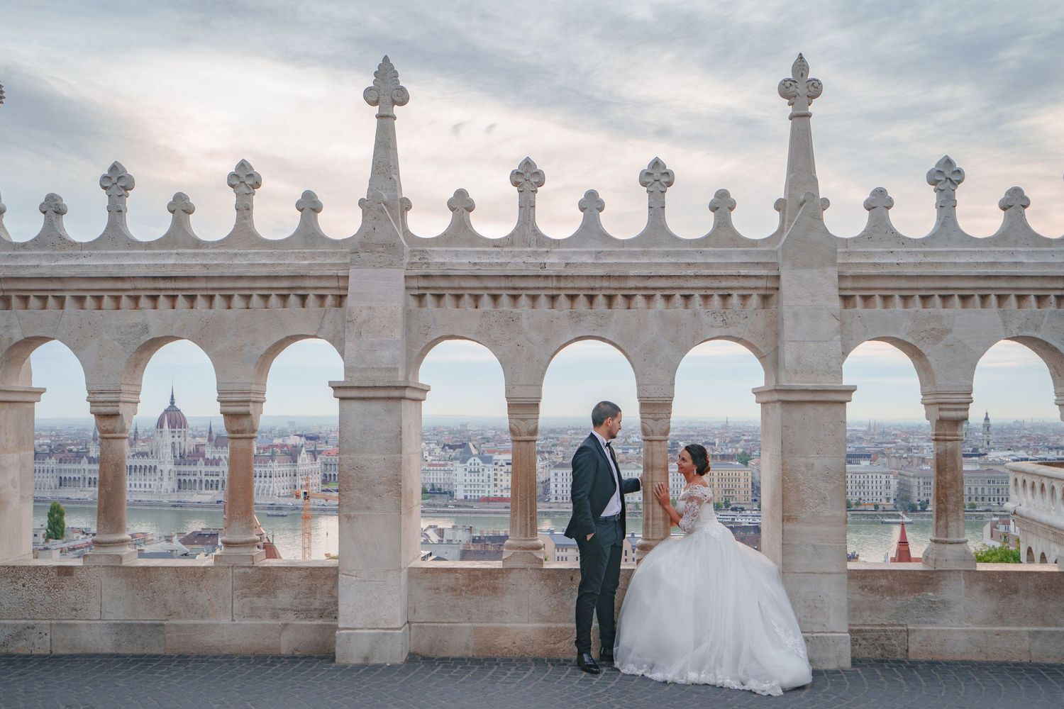 A couple in wedding attire poses against a gothic stone balcony overlooking a cityscape.