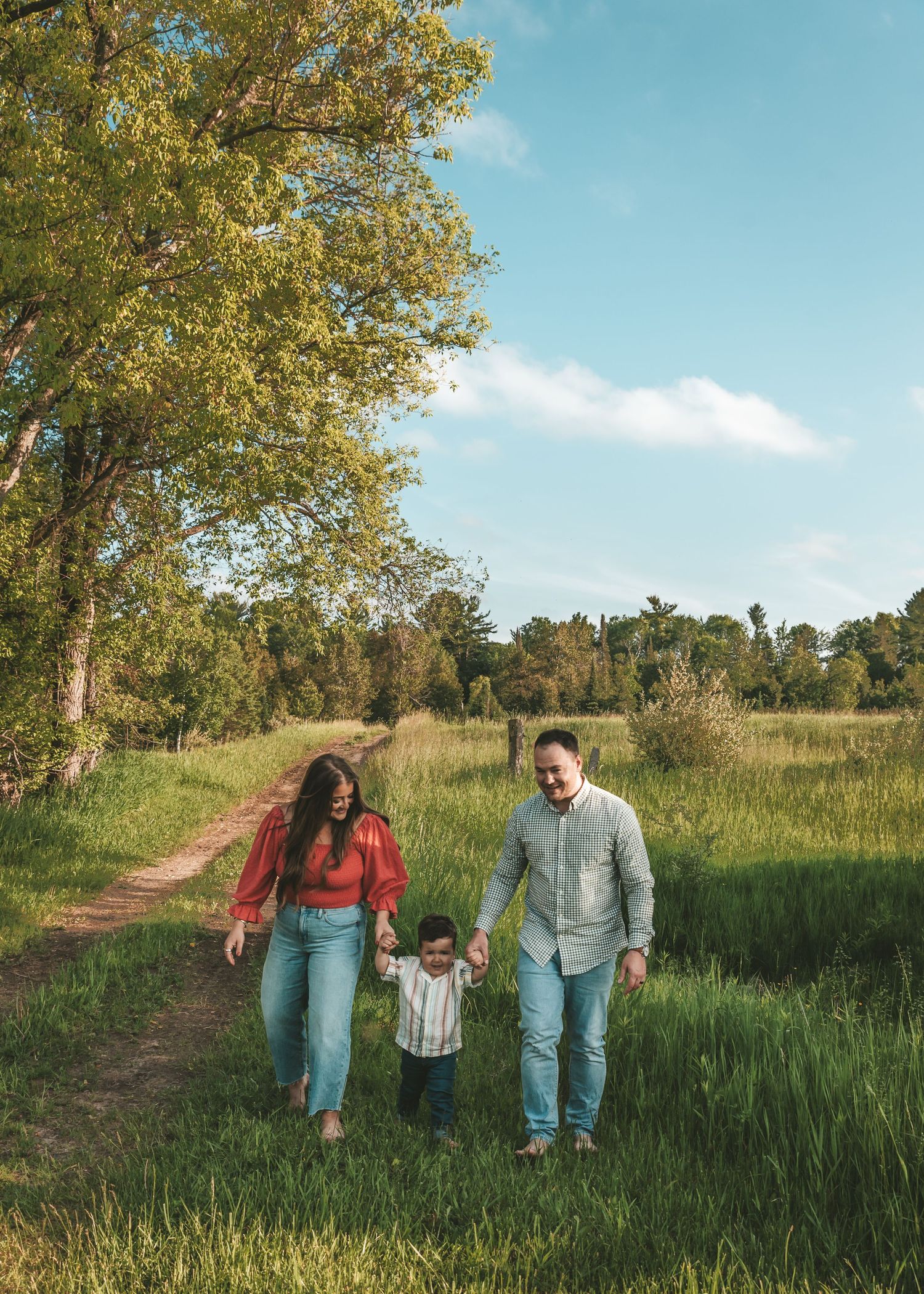 Family walking together on dirt path through sunny meadow with trees.