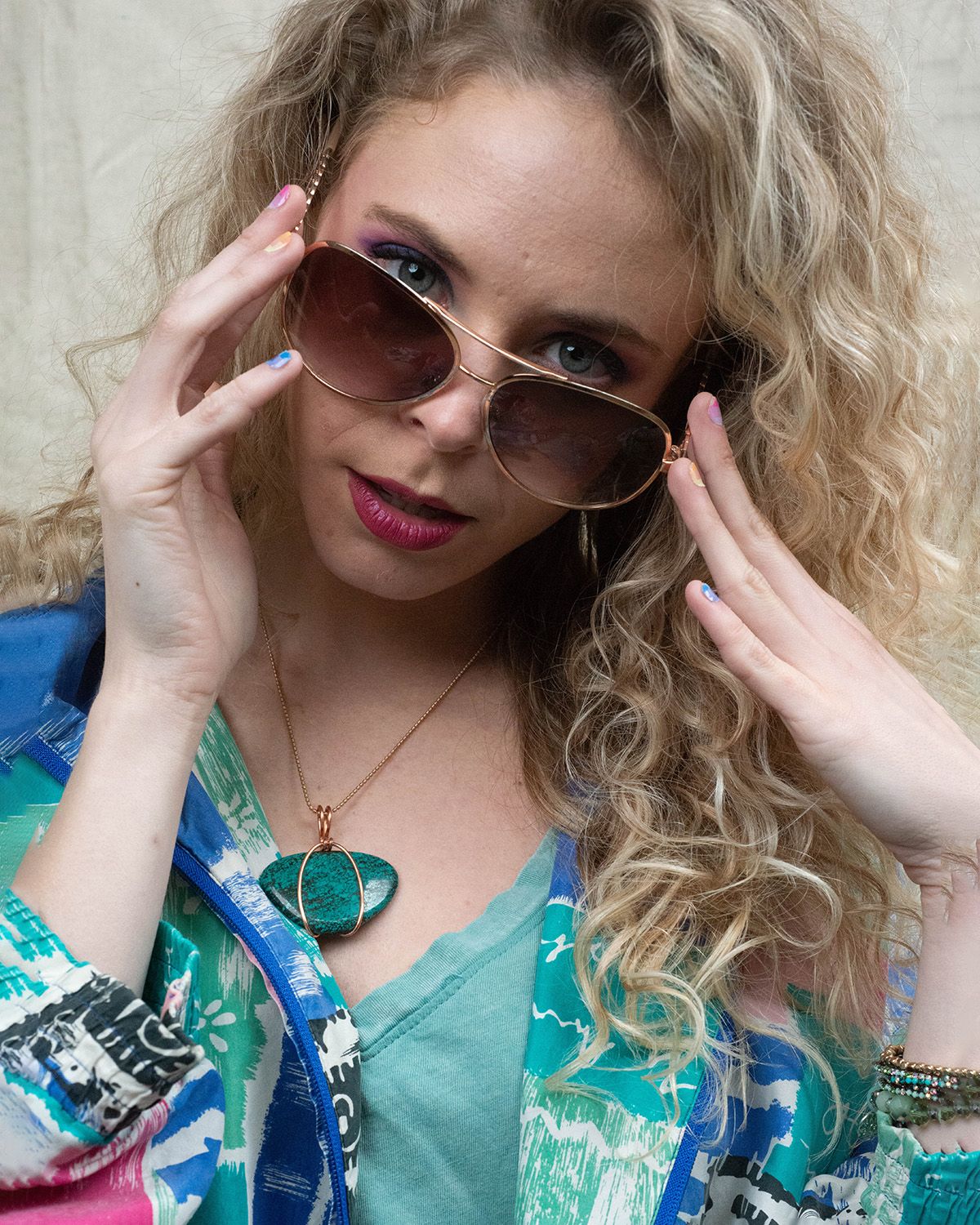 Person wearing sunglasses and a turquoise pendant poses in a blue and green patterned outfit with curly blonde hair.