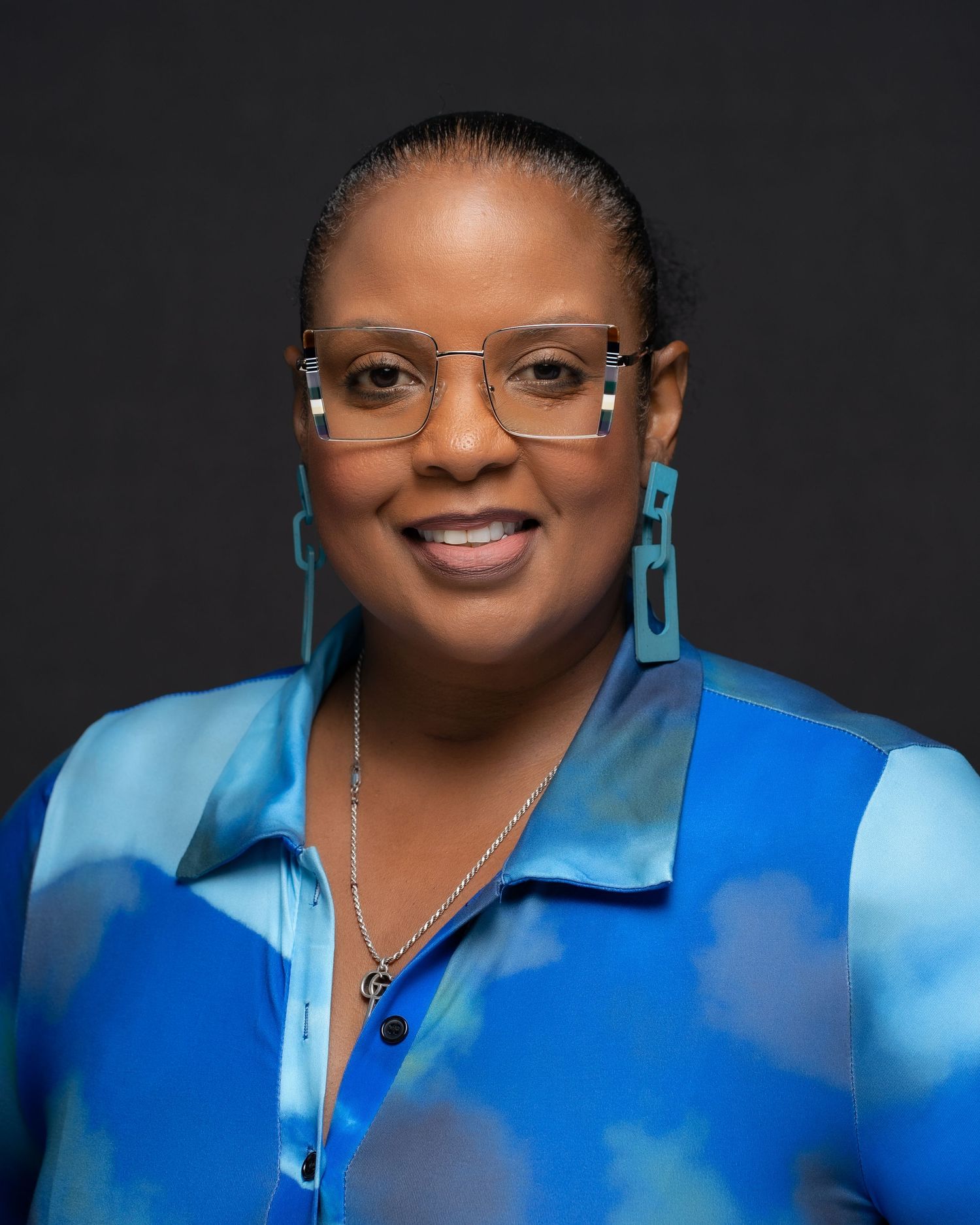 Smiling woman in a vibrant blue blouse with bold earrings and clear glasses, posing for a professional headshot