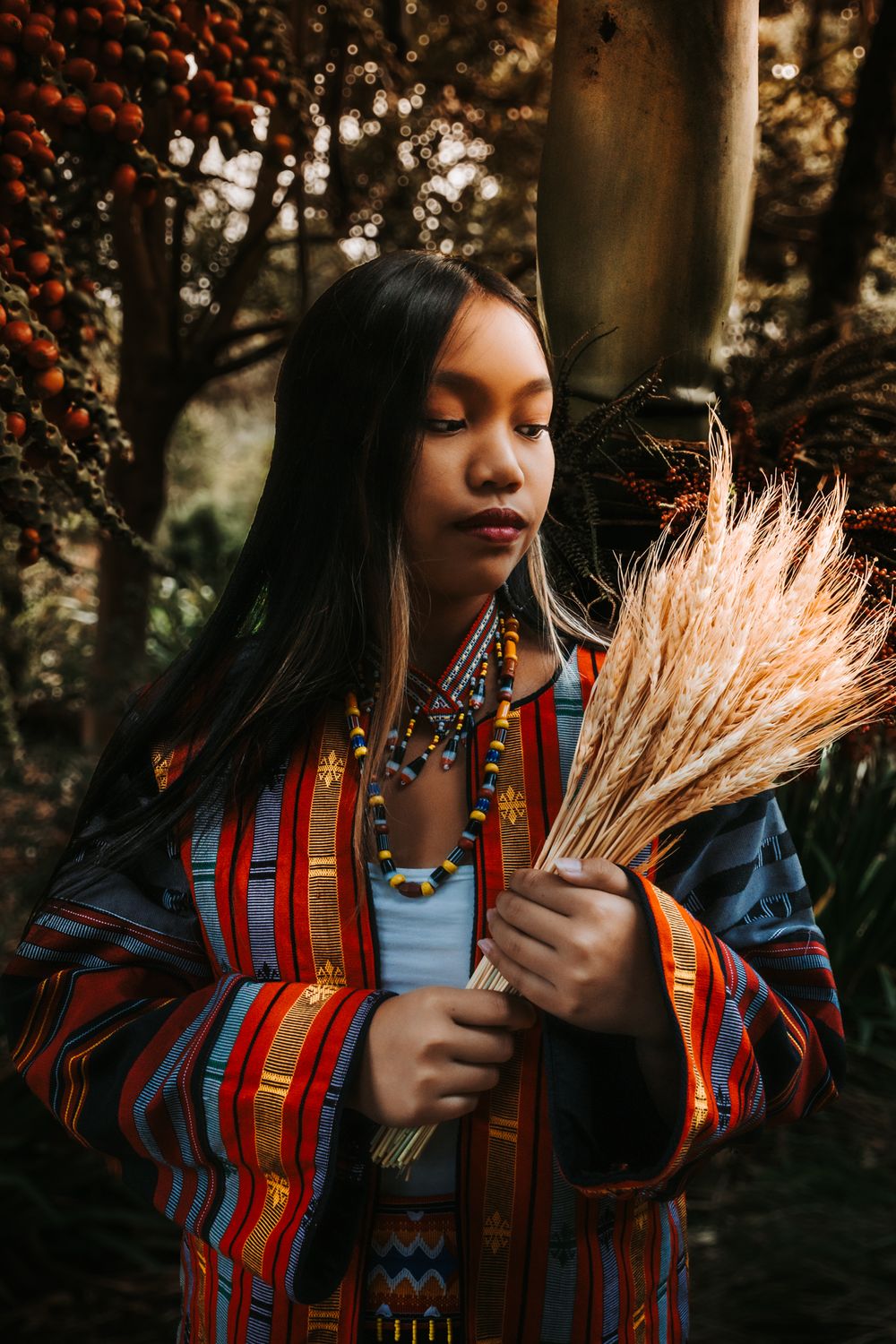Woman in colorful striped jacket holds dried wheat bundle in artistic outdoor portrait.
