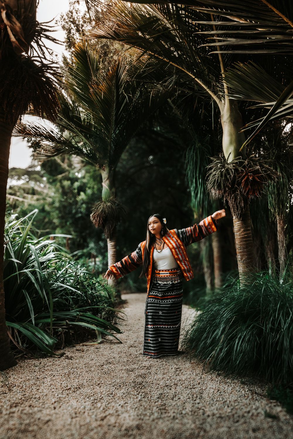 Model in patterned dress poses with outstretched arms on tropical garden path.