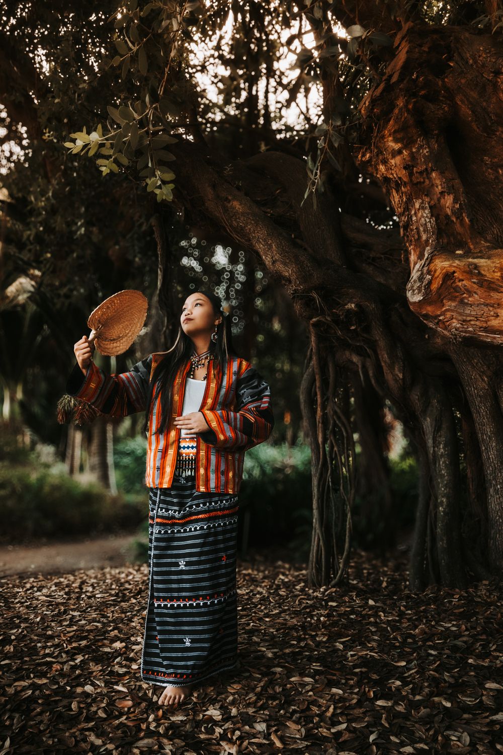 Person in traditional Igorot dress holds fan while looking up in forest setting.