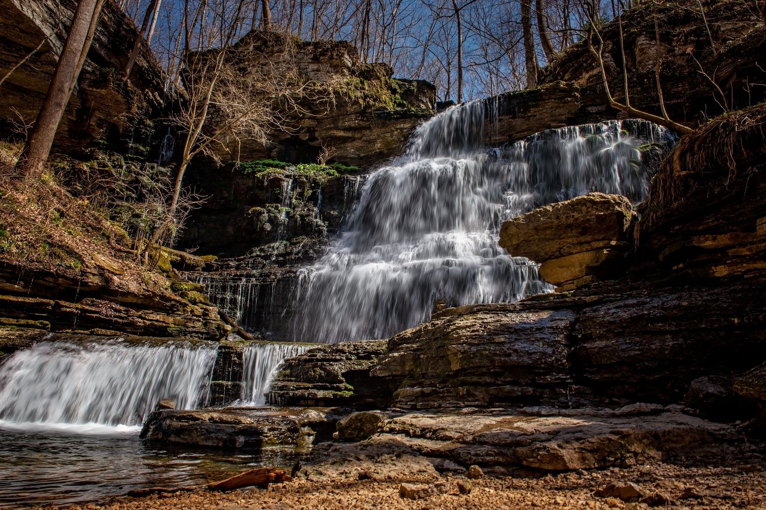Multi-tiered waterfall cascades over rocky ledges in a wooded forest setting.