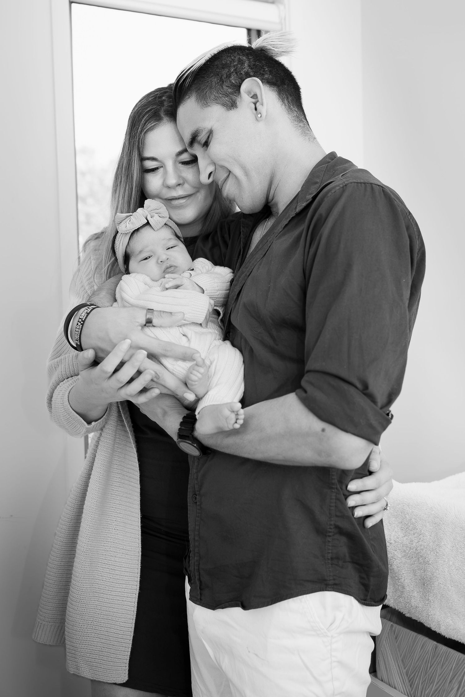 Black and white photo of two parents lovingly cradling their newborn baby girl wearing a bow headband.