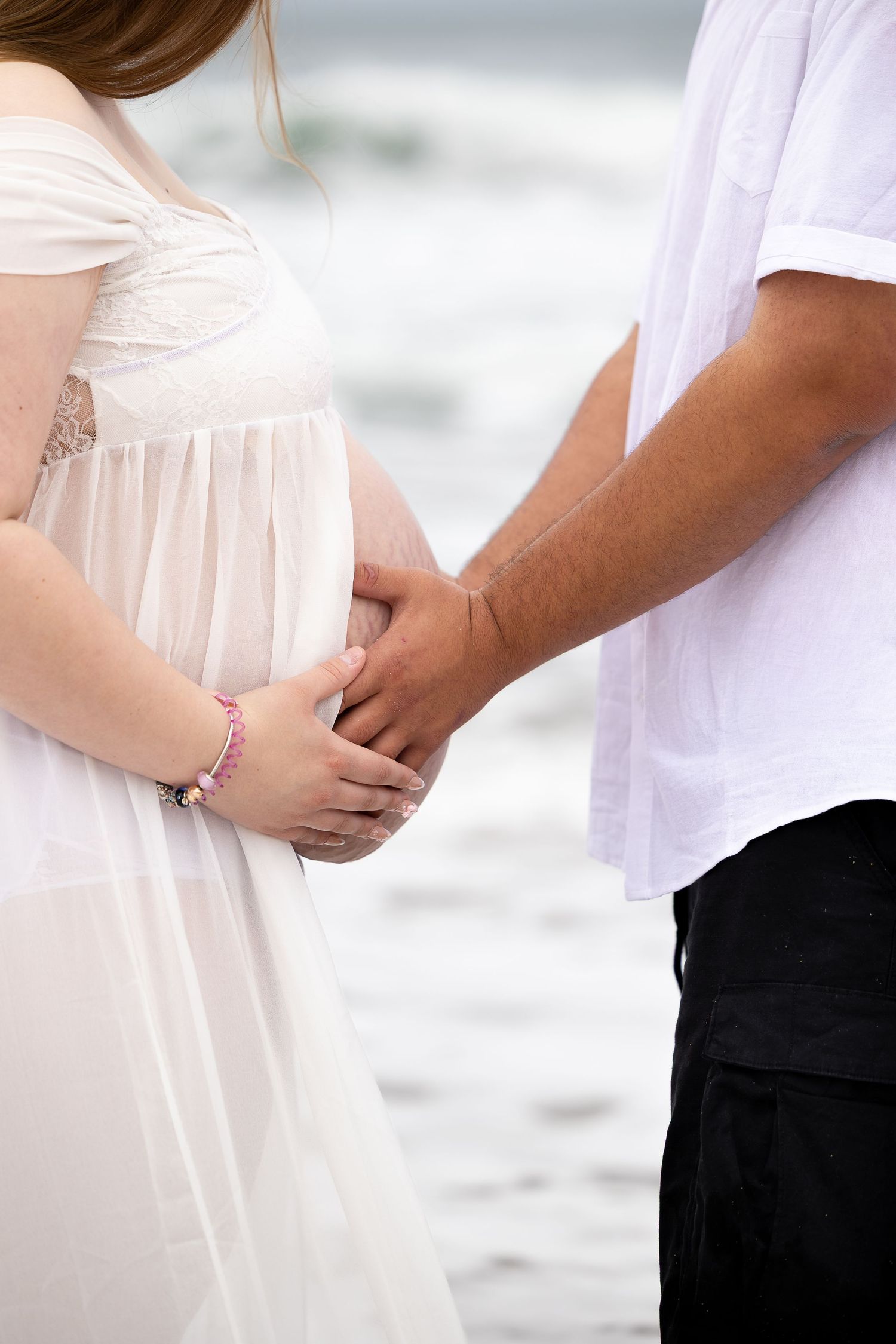 Pregnant woman in white dress and partner holding hands over baby bump at beach.