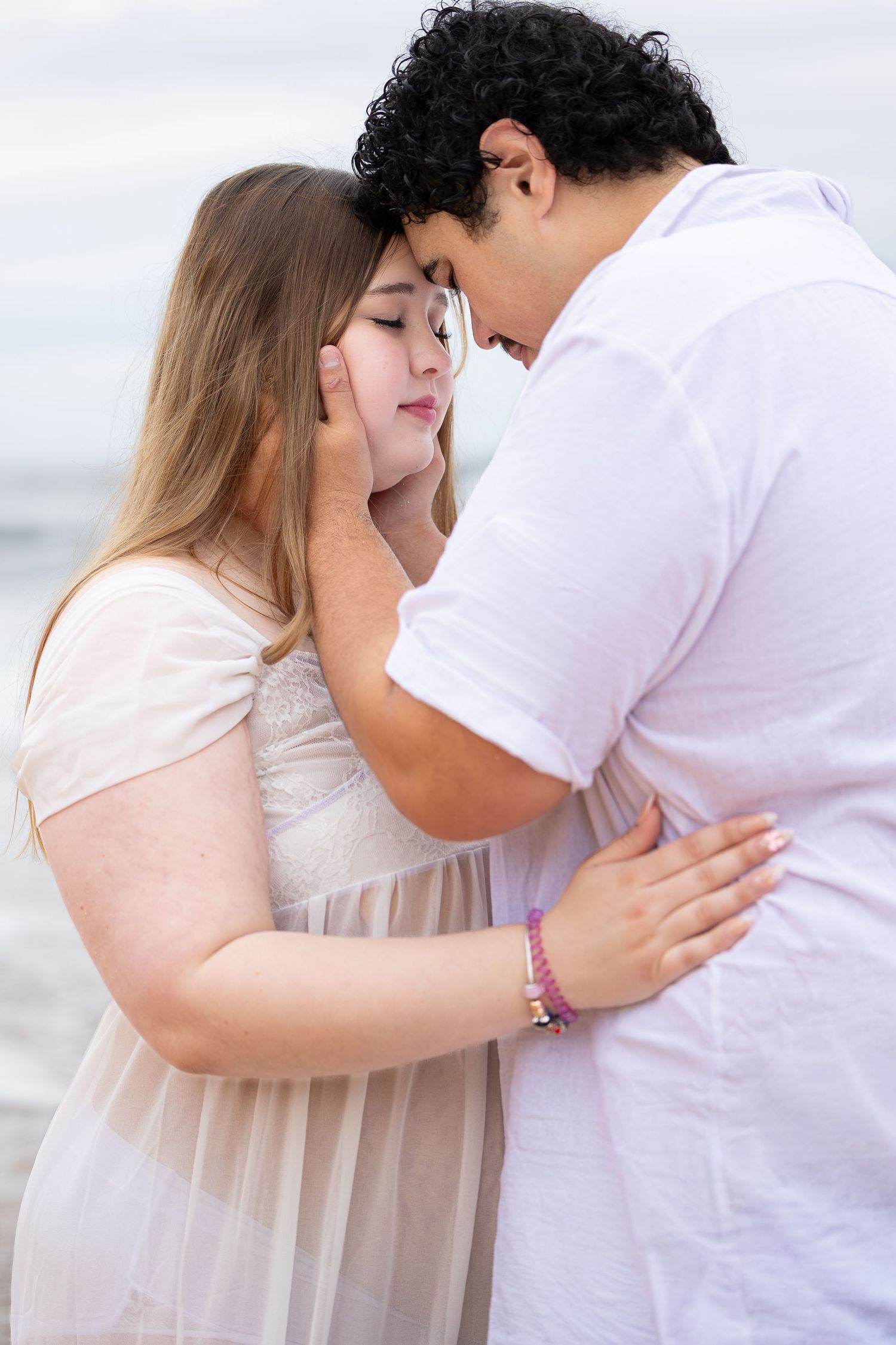 Romantic couple in white outfits sharing an intimate moment on a beach, foreheads touching tenderly.
