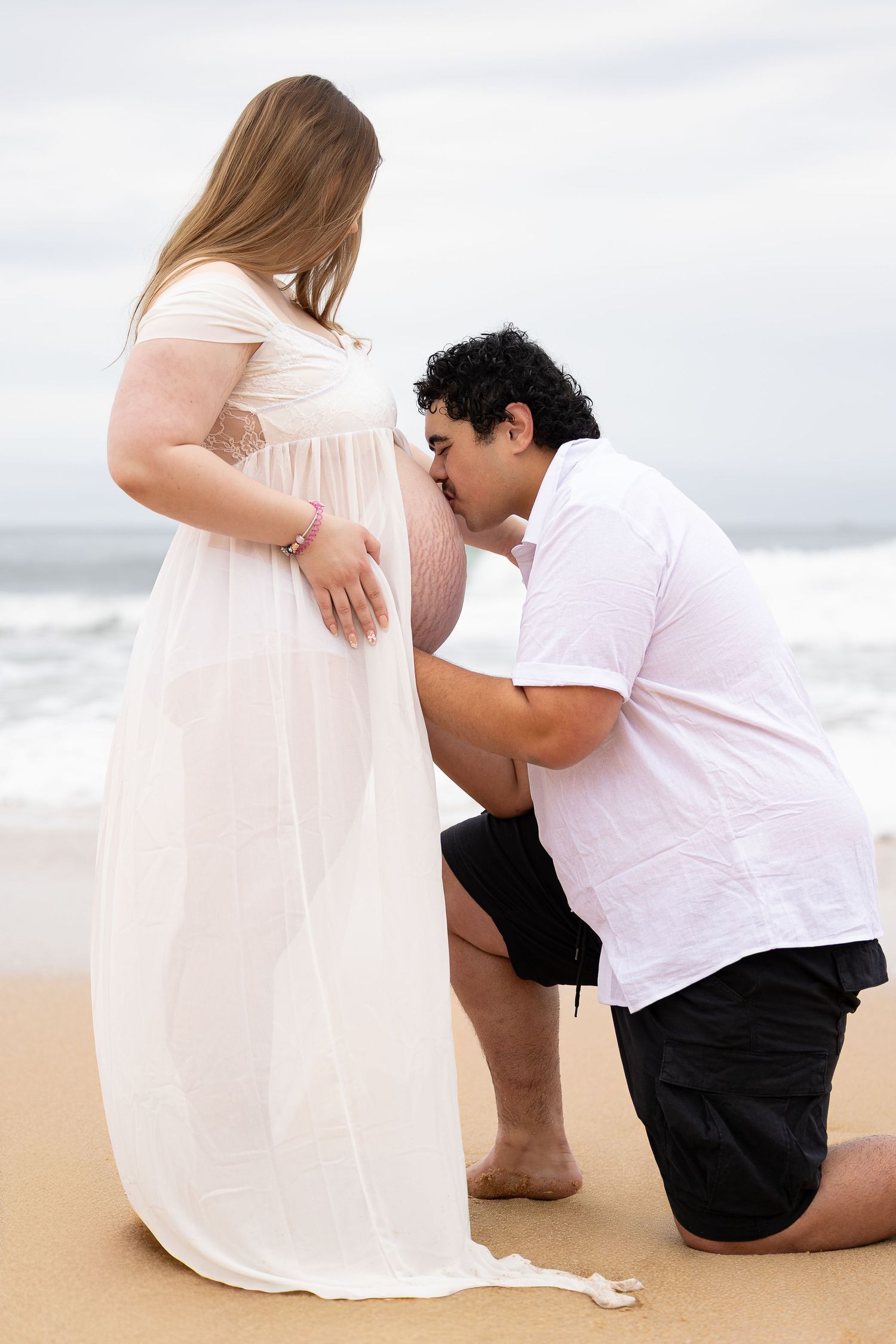 A man kneels on the beach kissing a pregnant woman's belly, both dressed in white and black near ocean waves.