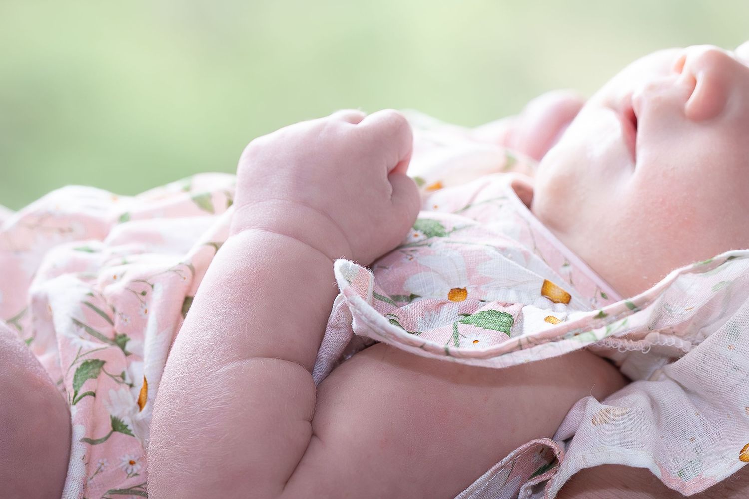 A baby wearing a pink and white floral dress lies on a soft surface while sleeping peacefully.