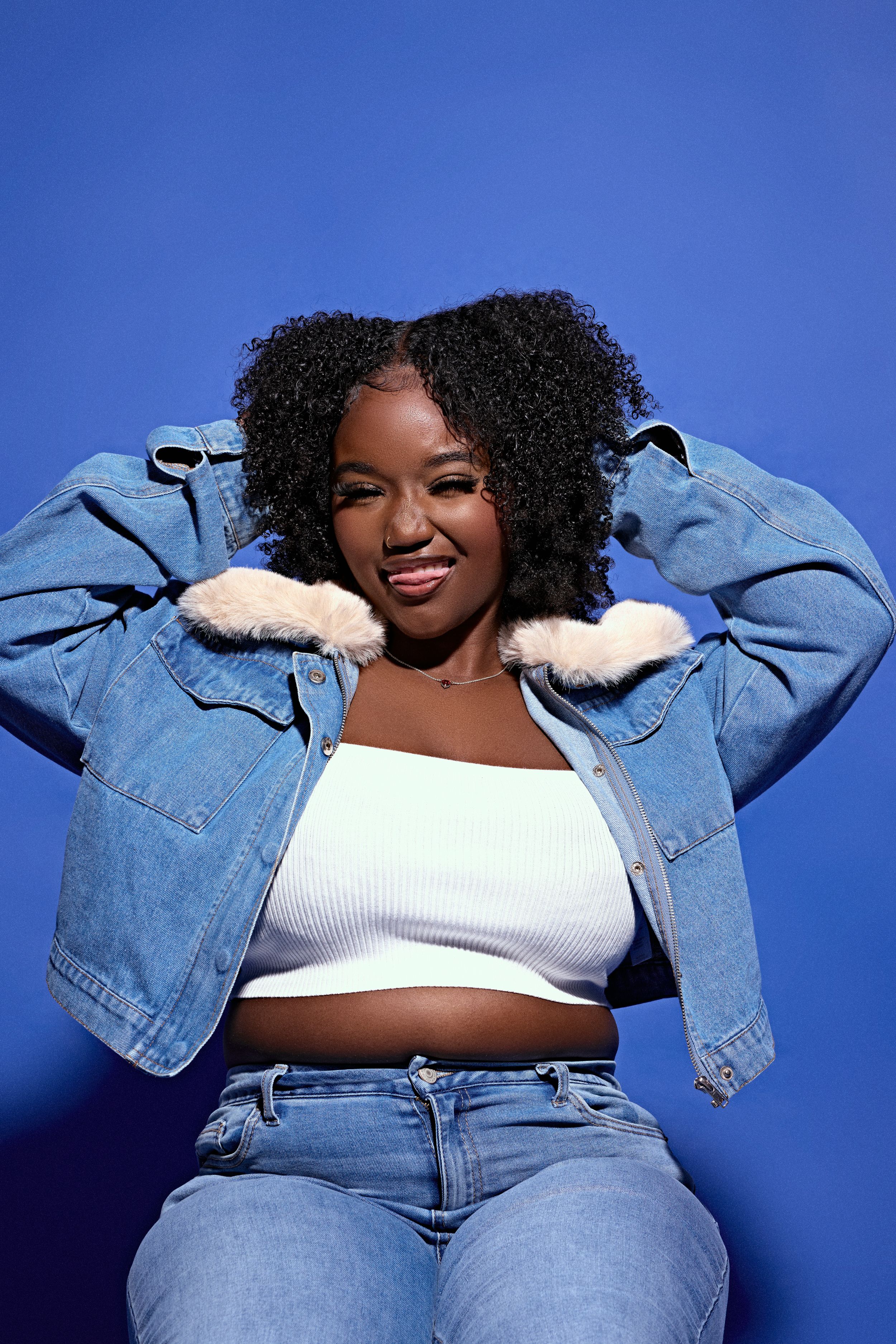 Portrait against blue background wearing white crop top and denim jacket with fur collar.