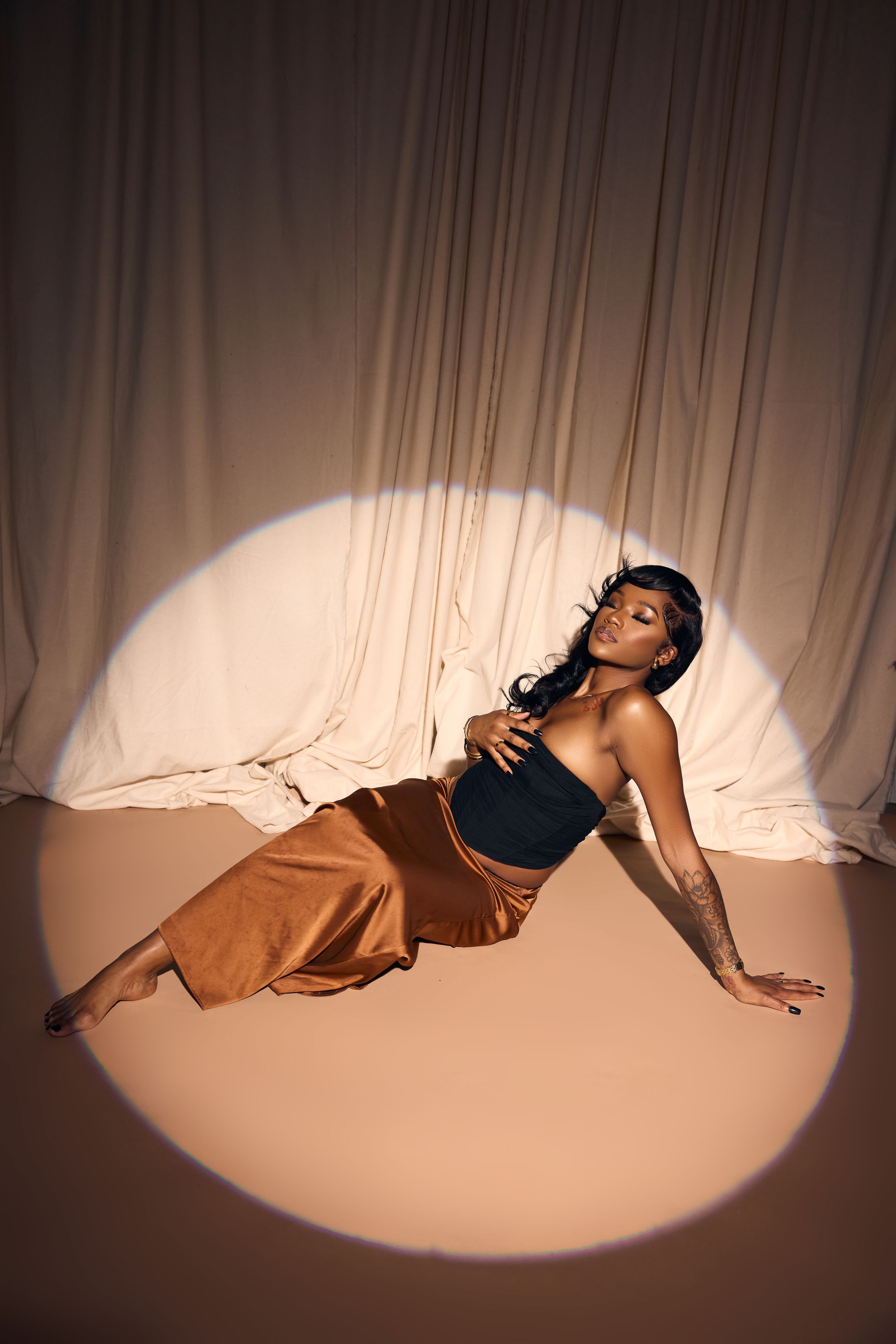Artistic fashion photo showing a model in a black top and brown flowy pants posing in a dramatic spotlight on light backdrop.