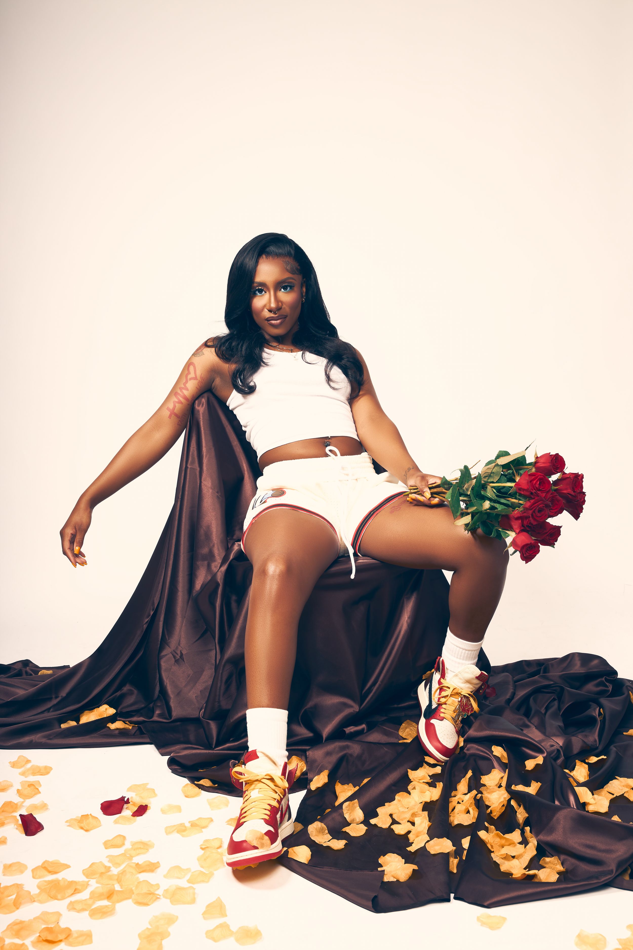 Fashion model in white athletic wear poses with red roses on brown silk fabric with scattered rose petals.