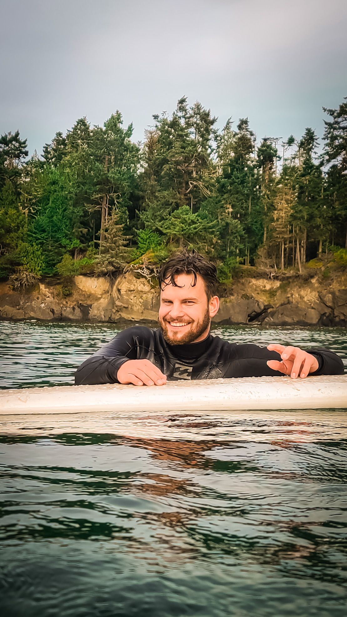 Person floating on a surfboard in calm waters near a forested shoreline.