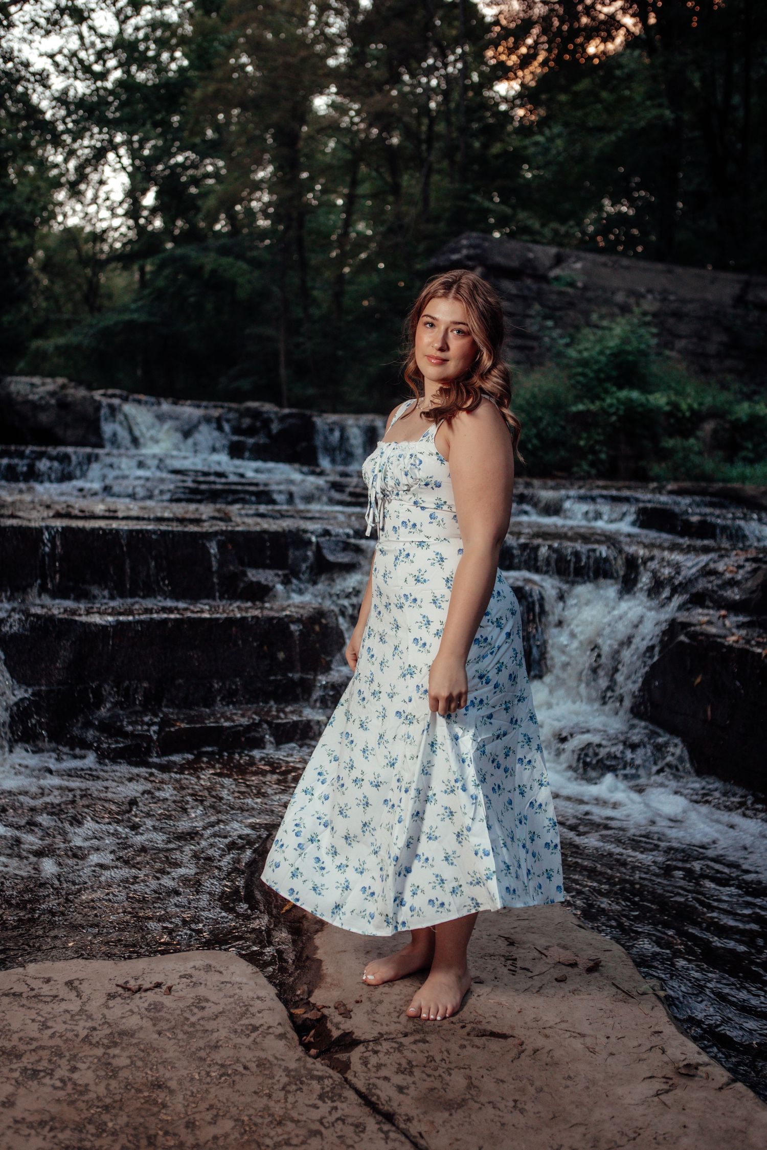A floral dress photoshoot by a cascading waterfall on rocky terrain at sunset.