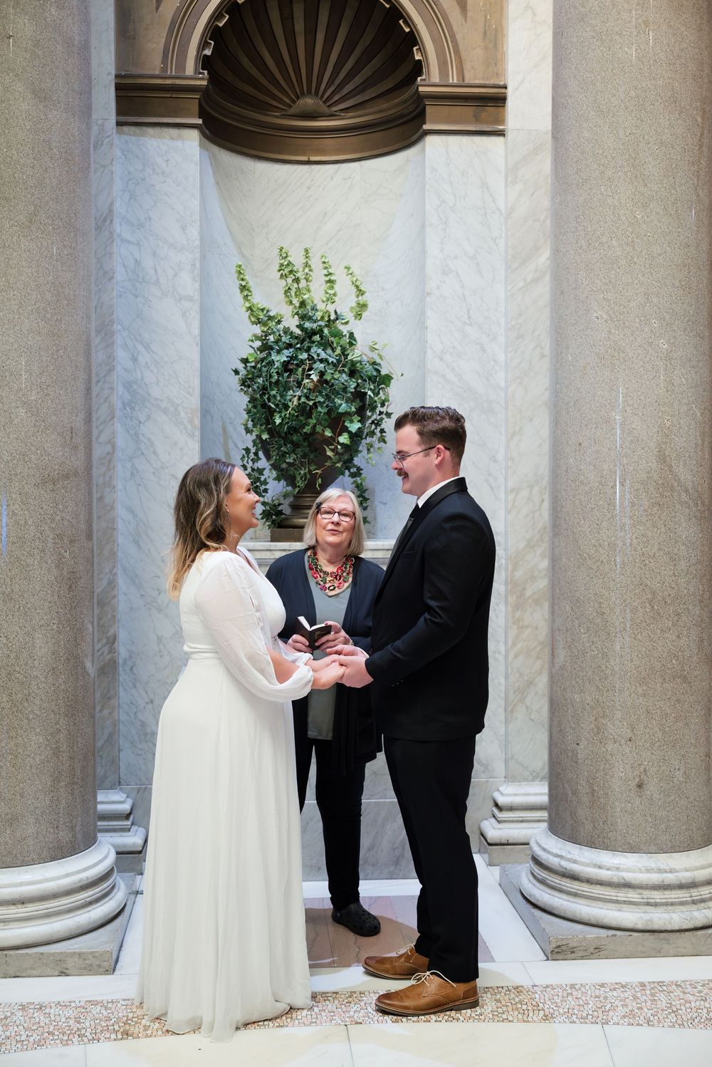 A wedding ceremony takes place between a couple in a classical columned alcove with an officiant.