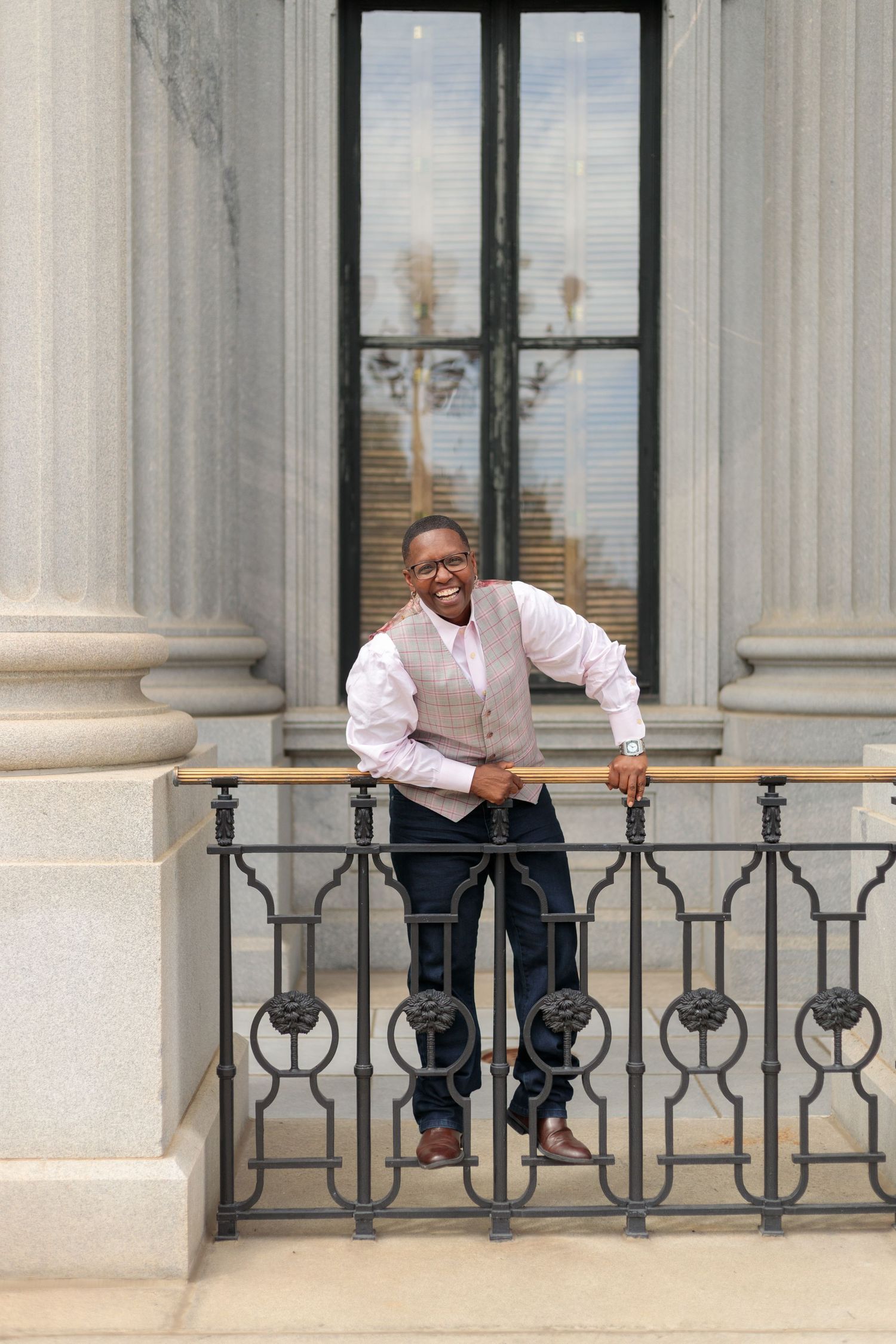 A person in business attire poses cheerfully on an ornate balcony with classical columns.