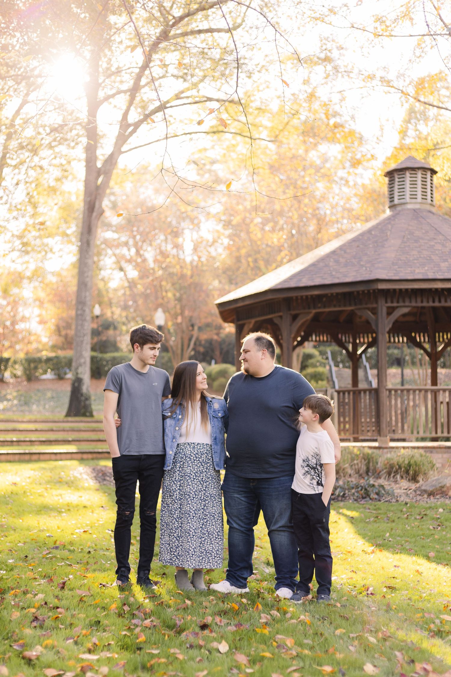 A family group stands together in front of a wooden gazebo on an autumn day with golden sunlight filtering through trees.