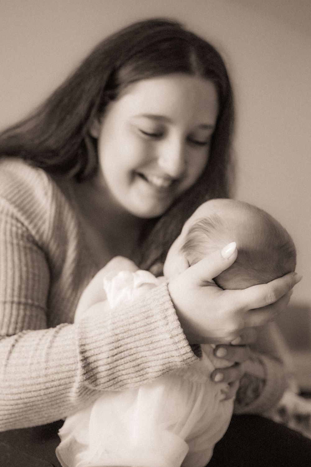 Smiling young woman in a knit sweater tenderly cradling a sleeping newborn baby in a sepia-toned portrait.