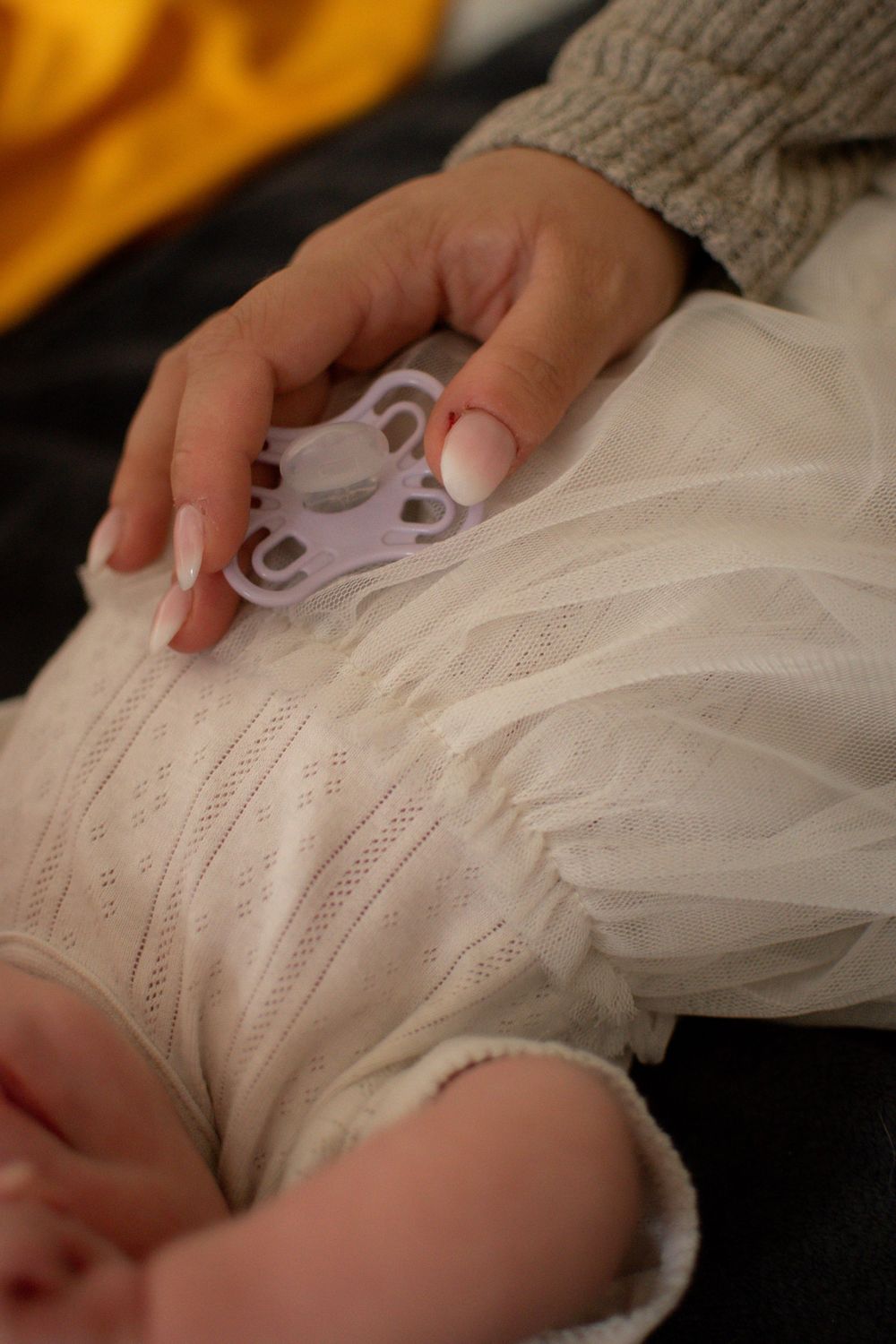 A parent's hand with pink nails holds a purple pacifier on a sleeping baby dressed in a white lace outfit.