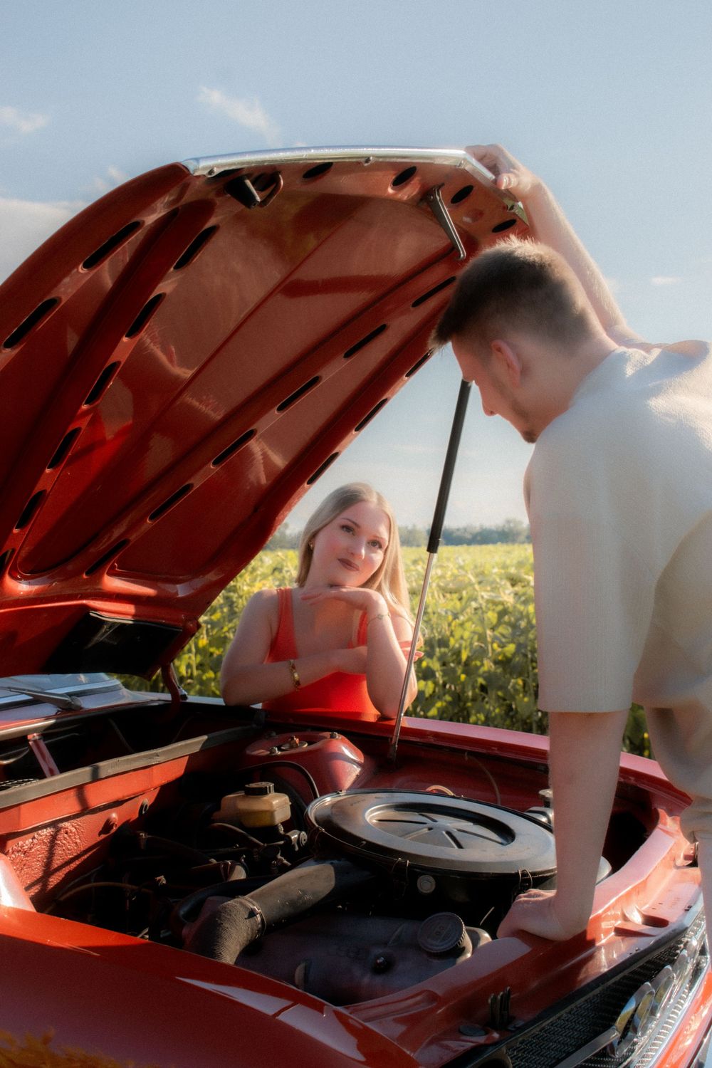 A young couple examines the engine of a red classic car in a sunny field, with the hood open.