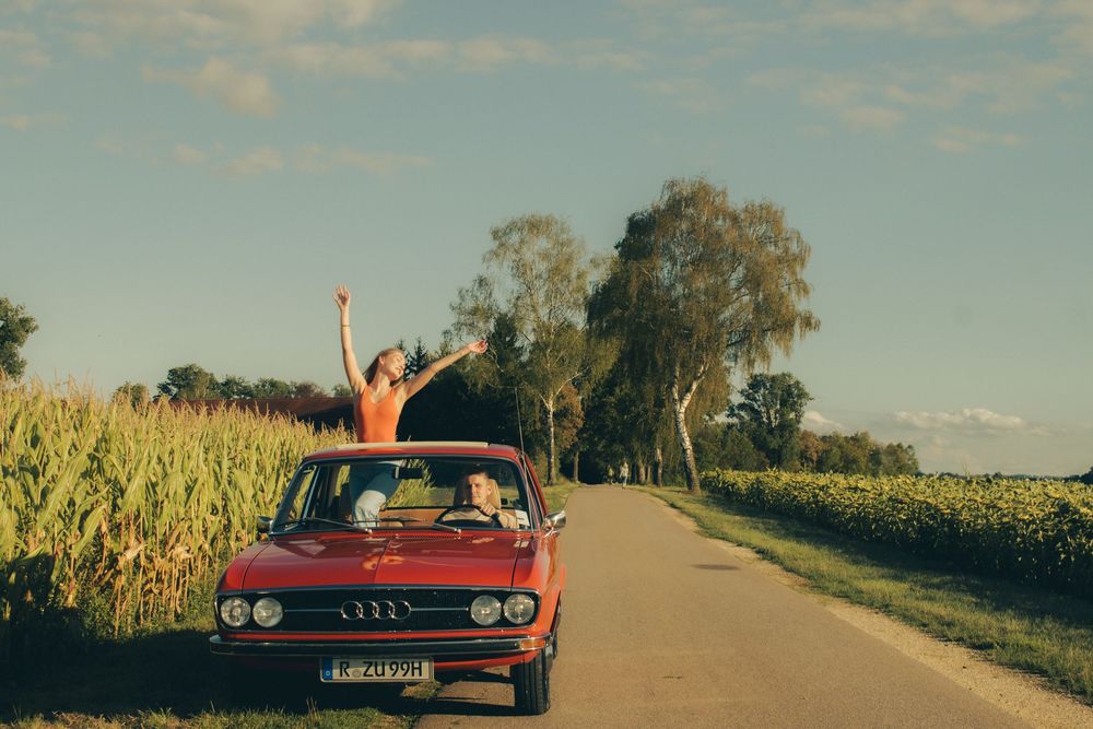 Two people joyfully riding in a vintage red Audi convertible on a rural road surrounded by cornfields.