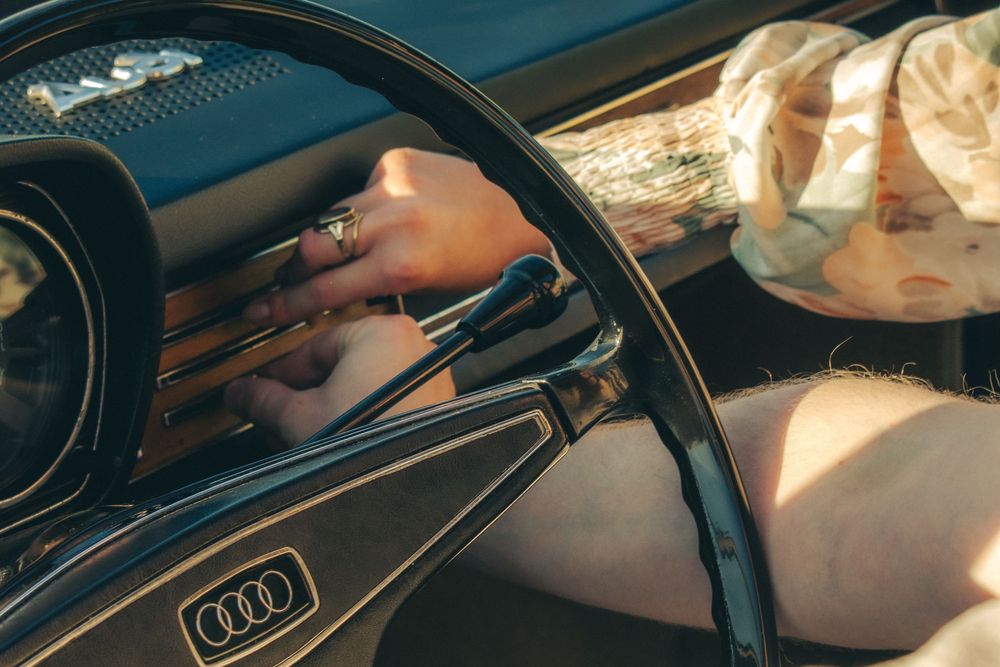 Close-up of a hand with a ring on the steering wheel of a luxury Audi car interior in warm sunlight.