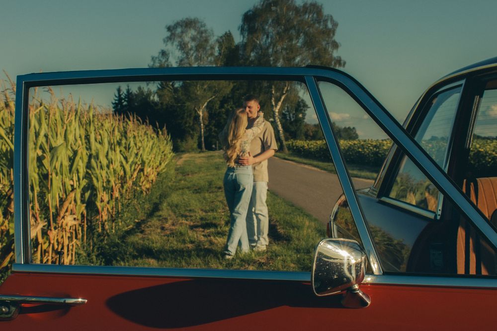 Couple embracing near a cornfield reflected in a vintage red car window on a sunny day.