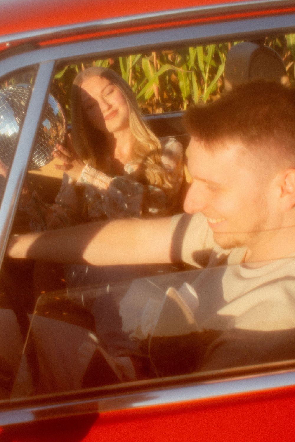 A smiling couple sitting inside a vintage red car on a sunny day, with green foliage visible in the background.