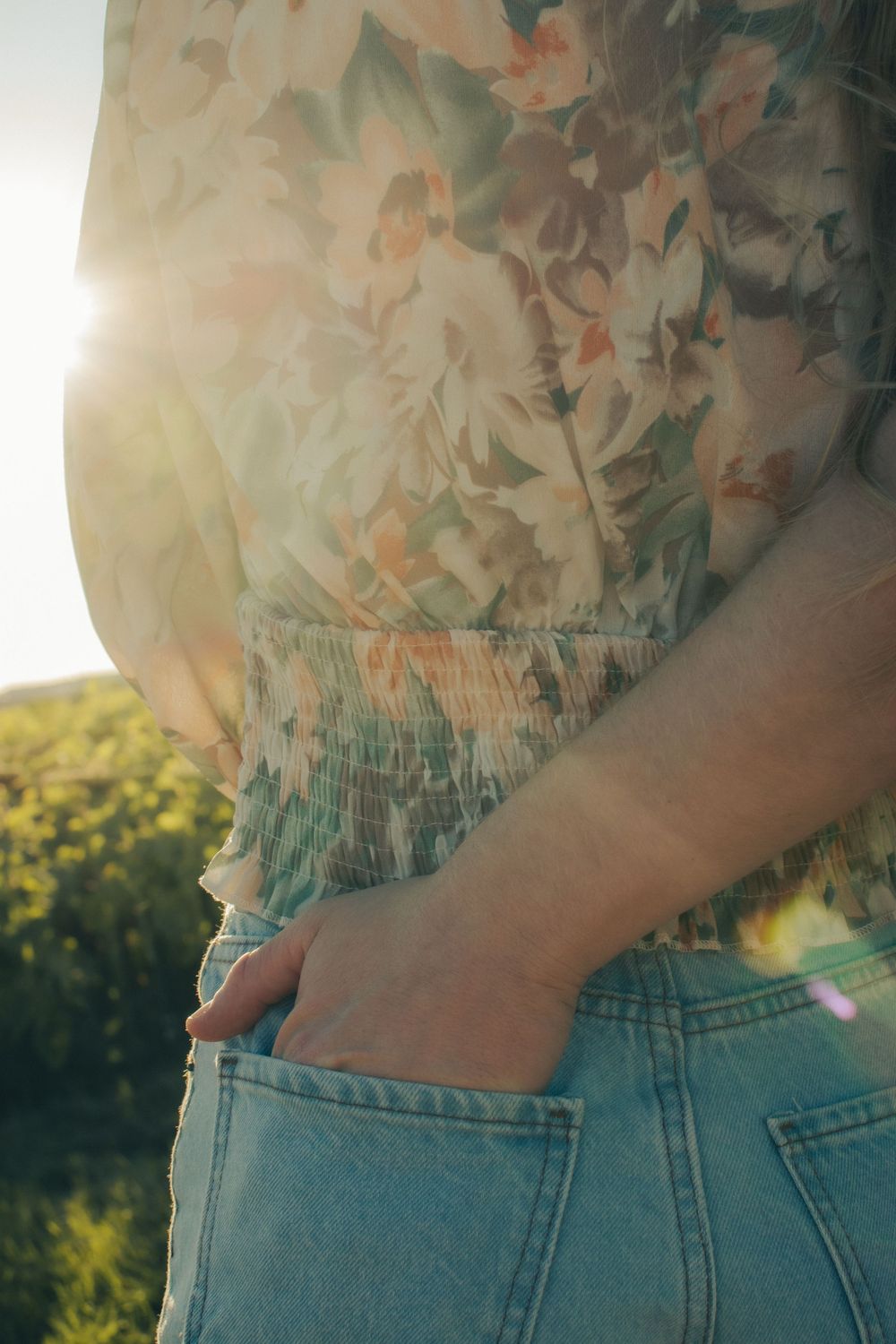 Woman in floral top and denim jeans with hand in pocket, standing in sunlit field.
